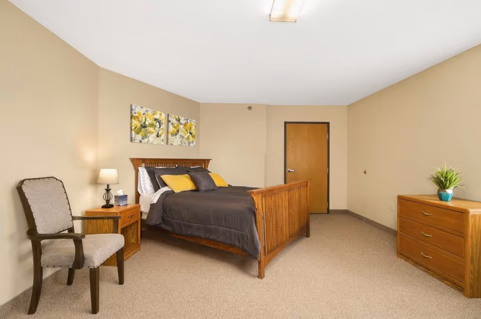 Furnished bedroom with a wooden bed, nightstand, chair and dresser in a neutral-toned room.