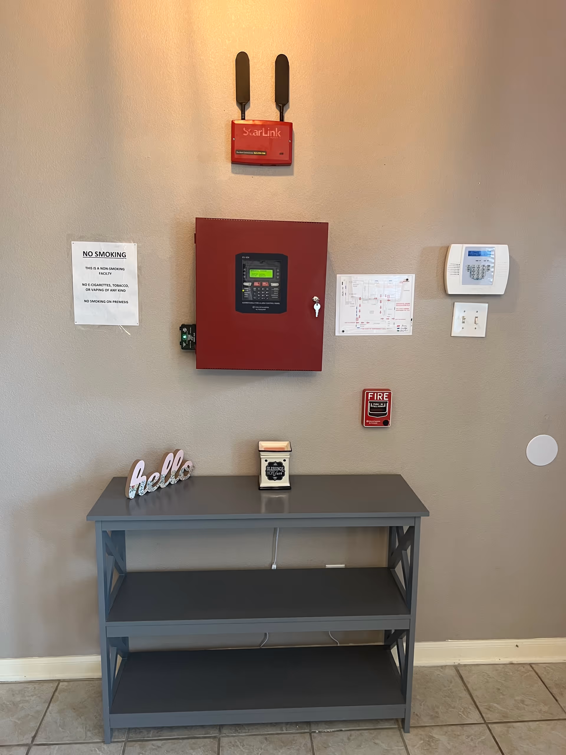 Gray console table with three shelves against beige wall, fire alarm panel and devices mounted above.