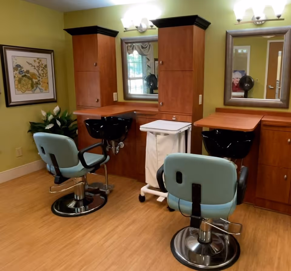 Interior view of a salon area in a senior living facility with two light blue salon chairs facing black wash basins. The room has wooden cabinets, two mirrors, a small rolling cart with a white laundry bag, a framed floral artwork on the wall, and a potted plant in the corner. The walls are painted green and the floor has a wood finish.