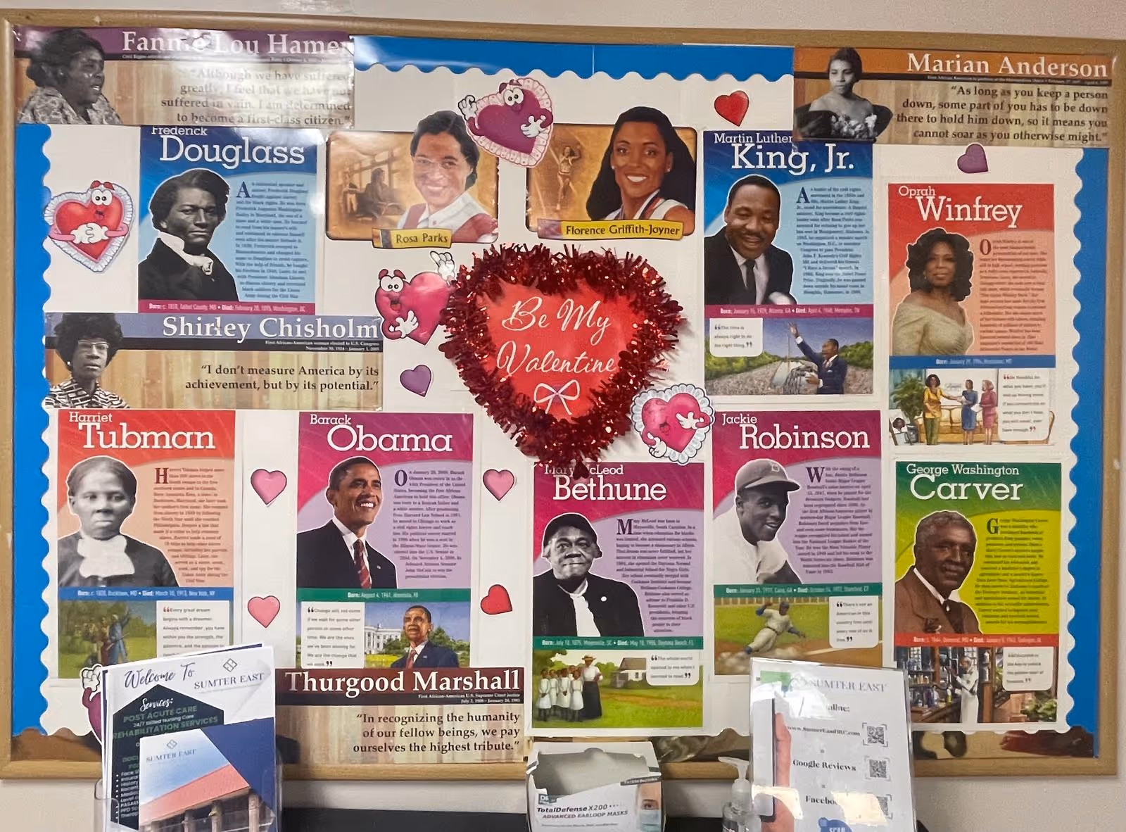 A bulletin board decorated with portraits and brief biographies of notable African American historical figures including Frederick Douglass, Rosa Parks, Martin Luther King Jr., Barack Obama, Harriet Tubman, Oprah Winfrey, and others. The board has a Valentine theme with heart decorations and a central red heart that says 'Be My Valentine.' Below the board are informational pamphlets and a box of face masks on a counter. The setting appears to be inside Sumter East Health and Rehabilitation Center.