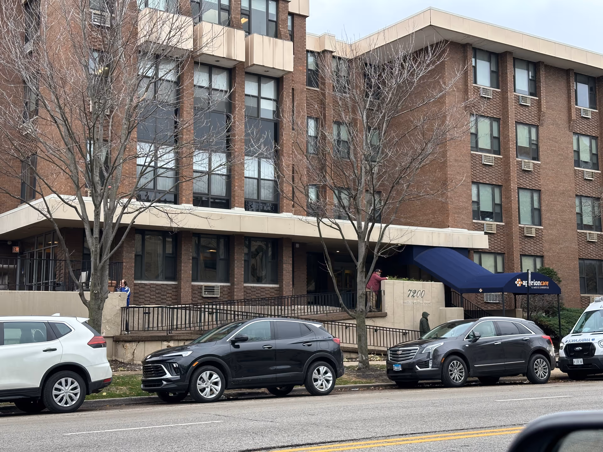 Exterior view of a multi-story brick building with several windows and air conditioning units. There is a blue awning over the entrance with the sign 'Aperion Care Lakeshore'. Several cars are parked along the street in front of the building, and a few leafless trees are visible on the sidewalk.