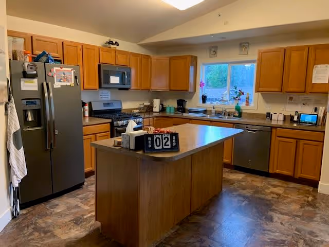 A spacious kitchen with wooden cabinets, a large island in the center, stainless steel refrigerator, dishwasher, and microwave. The kitchen has a window above the sink with plants on the windowsill and various kitchen appliances on the countertops.