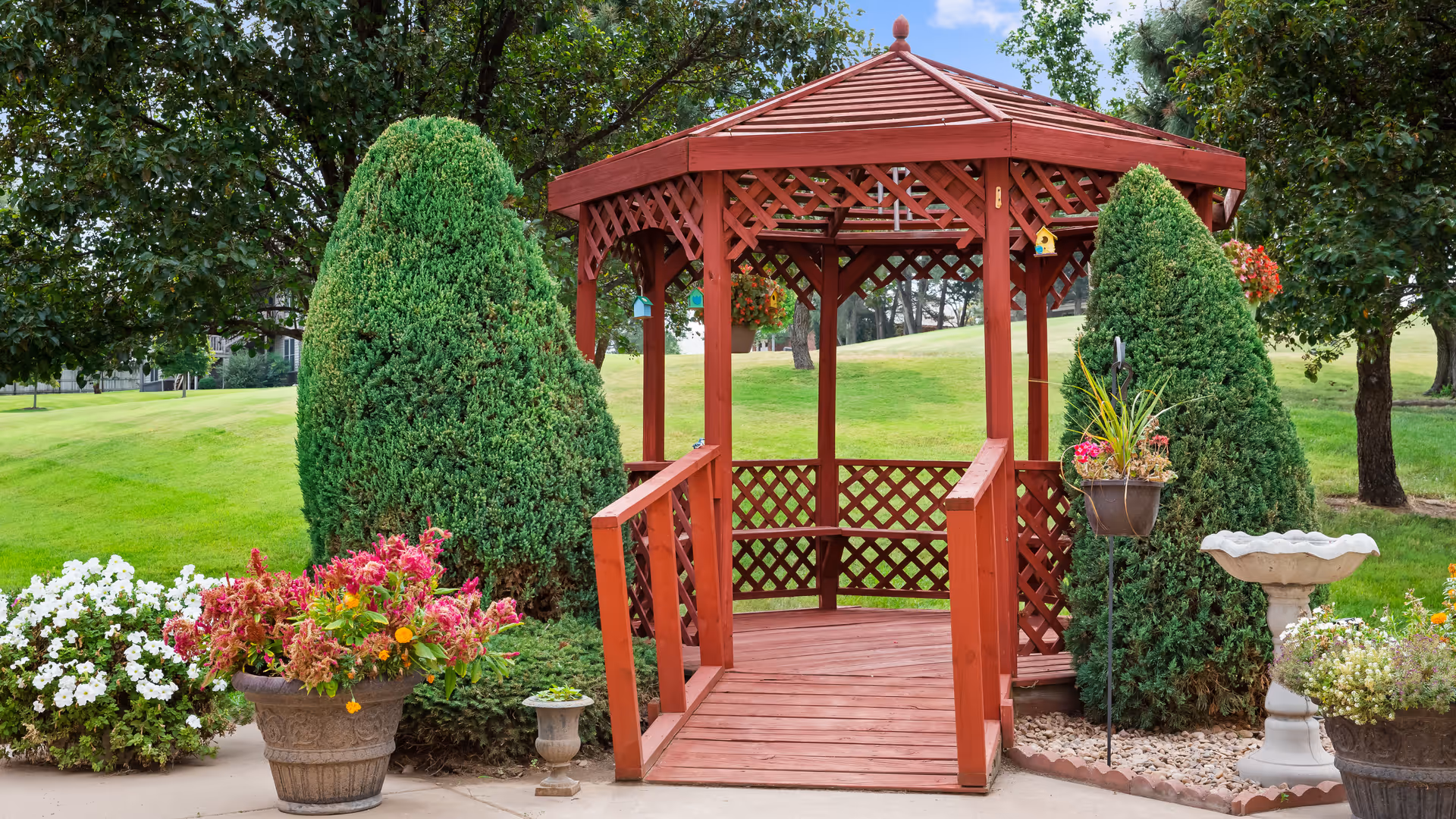 A red wooden gazebo with lattice sides and a small ramp entrance, surrounded by green grass, trimmed bushes, and colorful flower pots in a garden setting.