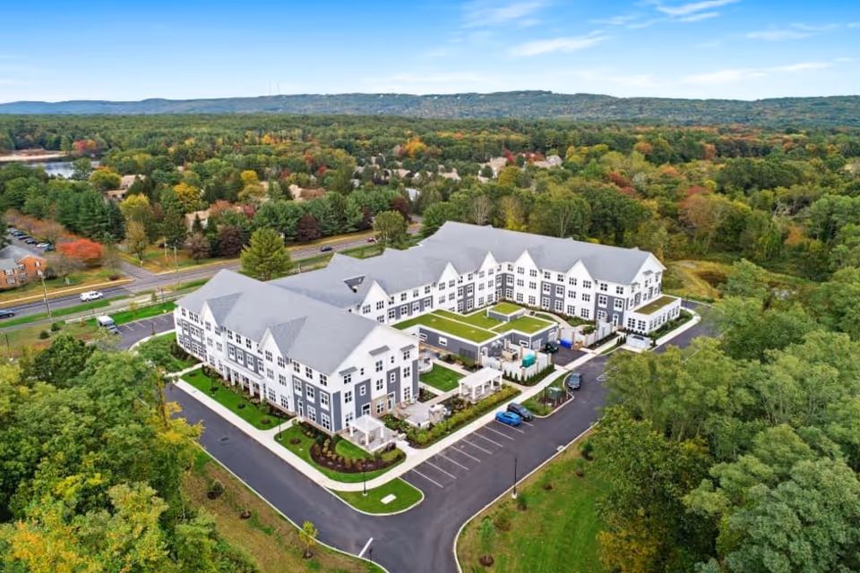 Aerial view of a large senior living facility named Alto Overland Park surrounded by trees and greenery with a parking lot and roads nearby. The building has a modern design with multiple peaked roofs and outdoor seating areas.