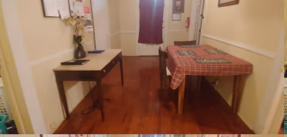 Interior view of a room with wooden flooring, a table covered with a red plaid tablecloth and placemats, and a small side table with a vase holding dried flowers. The walls are painted light yellow with white trim, and there is a door at the far end with a dark curtain.