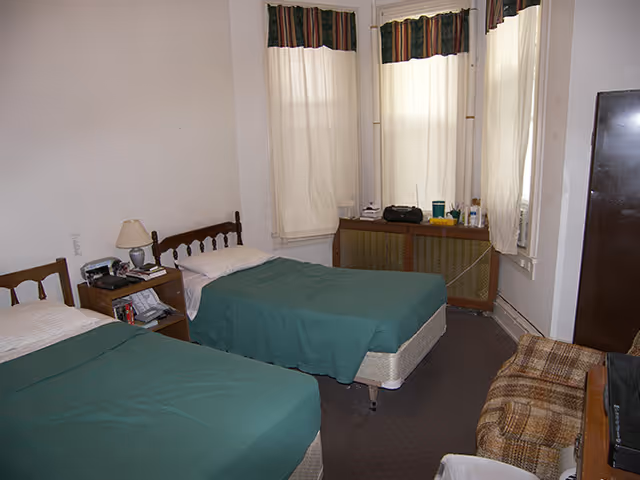 A simple bedroom with two twin beds covered in green bedspreads, a wooden nightstand with a lamp and some items, three windows with white curtains and valances, a brown plaid armchair, and a dark wardrobe.