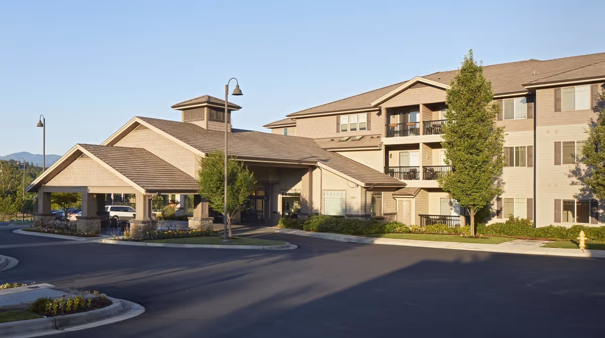 Exterior view of a three-story senior living building with a covered porte-cochère entrance, balconies, and landscaped driveway.
