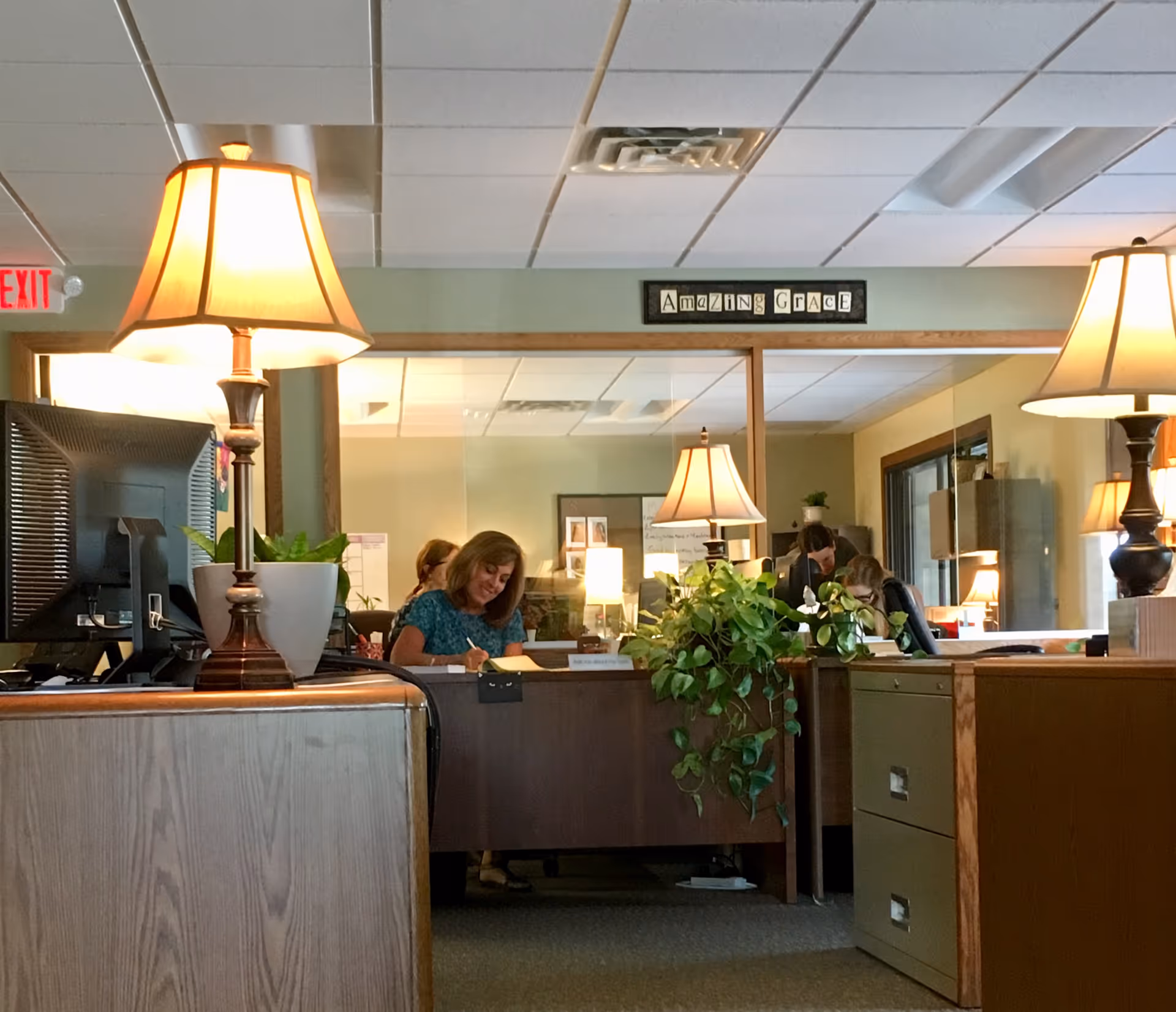 Reception area with staff at desks, lamps, plants, and a sign reading Amazing Grace.