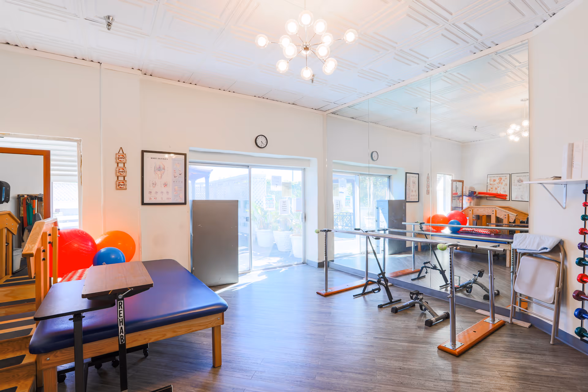 Bright rehabilitation room with a padded therapy table labeled 'REHAB', colorful exercise balls, parallel bars for walking practice, a large wall mirror, and anatomical charts on the walls. The room has wooden flooring, a white ceiling with a modern chandelier, and sliding glass doors leading outside.