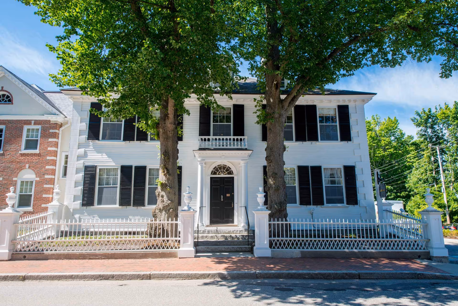 White two-story building with black shutters, ornate entryway, and large trees in front.