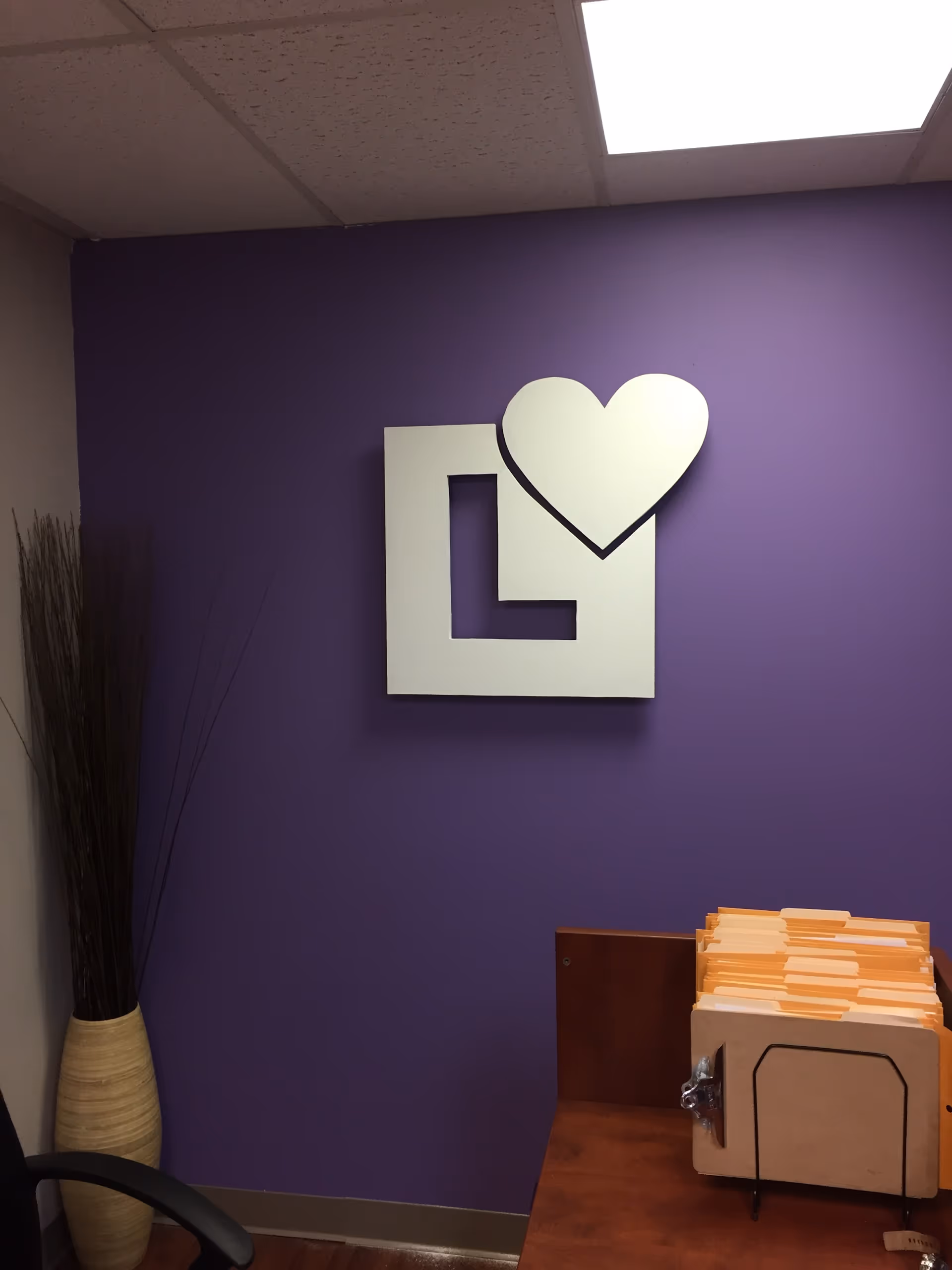 Purple wall with a white L heart logo above a desk with orange folders.