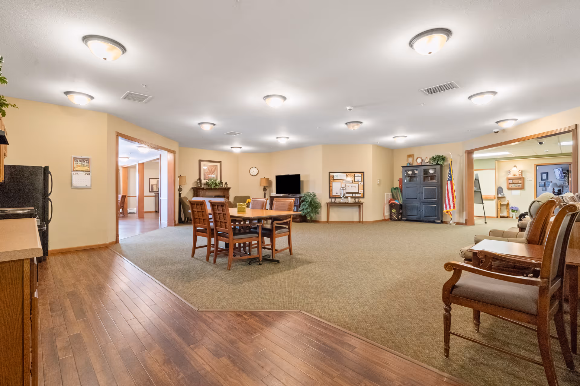 Spacious senior living common room with a central dining table, multiple chairs, a TV, and cabinetry.