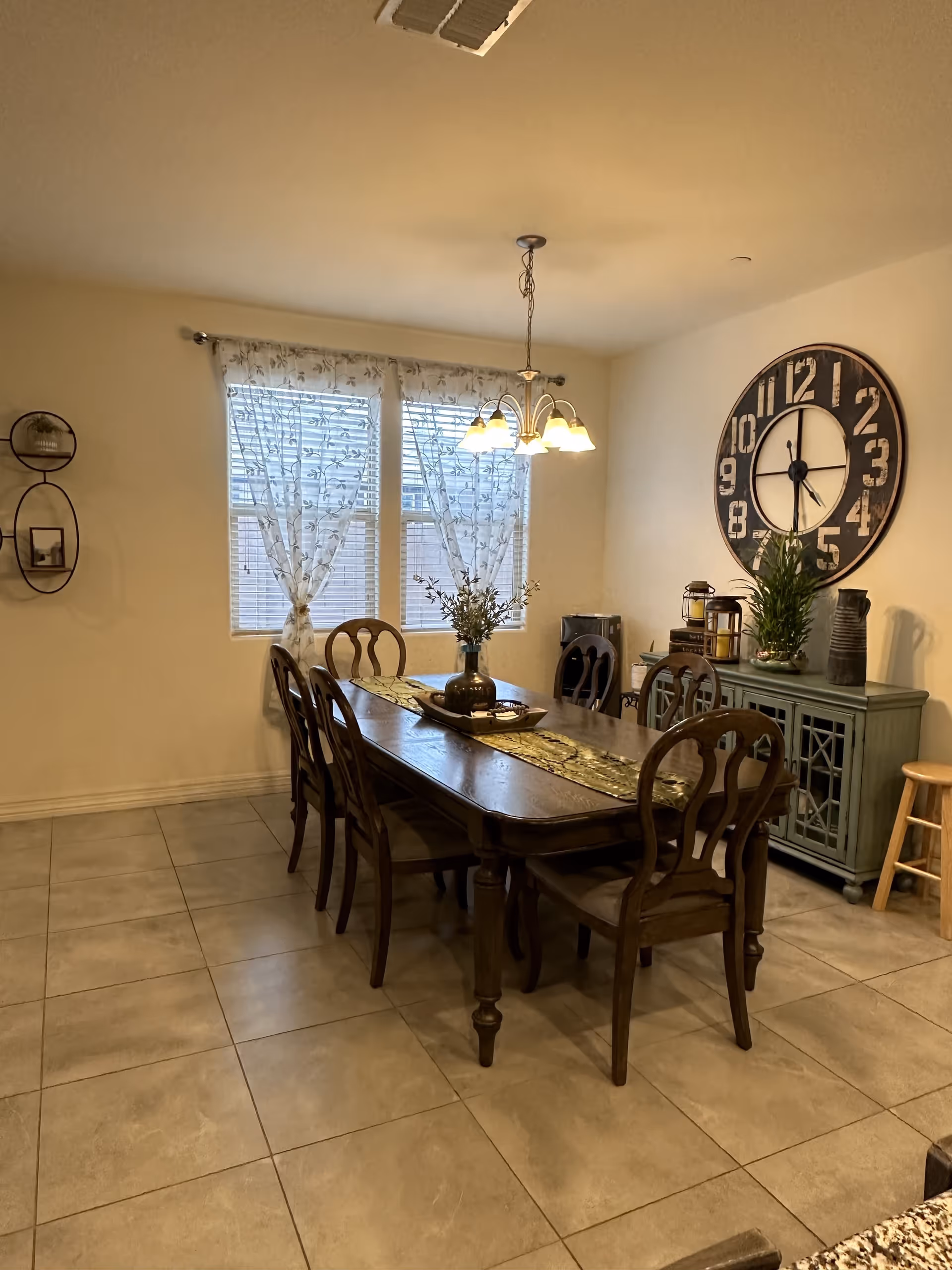 Dining room with a long wooden table, chandelier, windows, and a large wall clock.