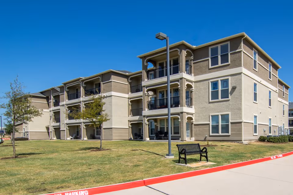 Three-story beige apartment building with covered balconies, a bench and lamp post on a grassy lawn under a clear blue sky.