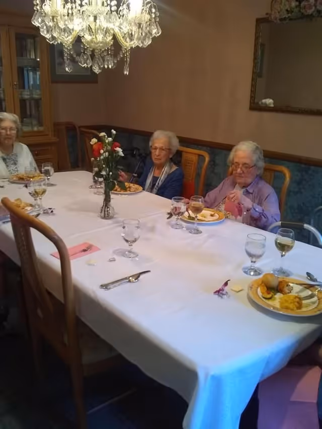 Three elderly women sitting at a dining table with a white tablecloth, eating a meal. The table has plates with food, glasses of water and wine, and a vase with flowers in the center. The room has a chandelier, wooden chairs, and a cabinet in the background.