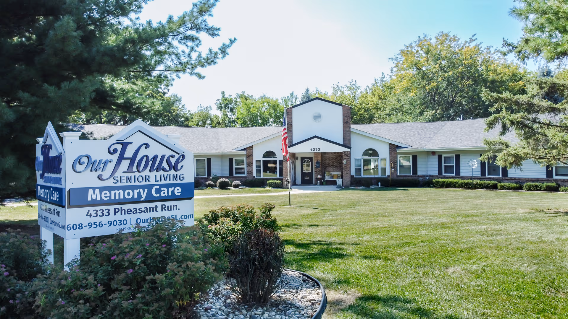 Front view of a single-story senior living memory care building with a lawn and a large sign reading 'Our House Senior Living Memory Care'.