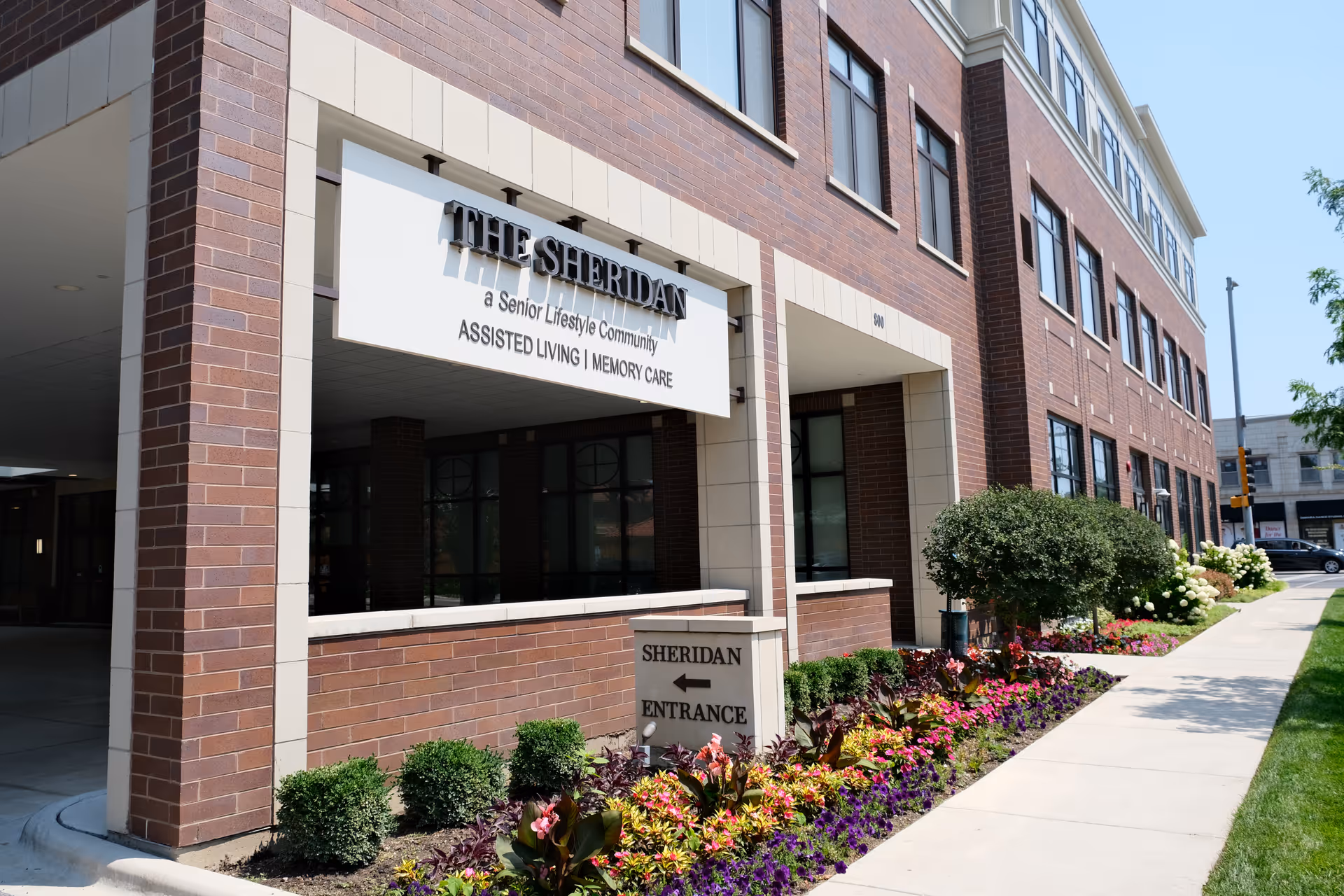Brick exterior and landscaped sidewalk leading to the entrance of The Sheridan assisted living building with a canopy sign.