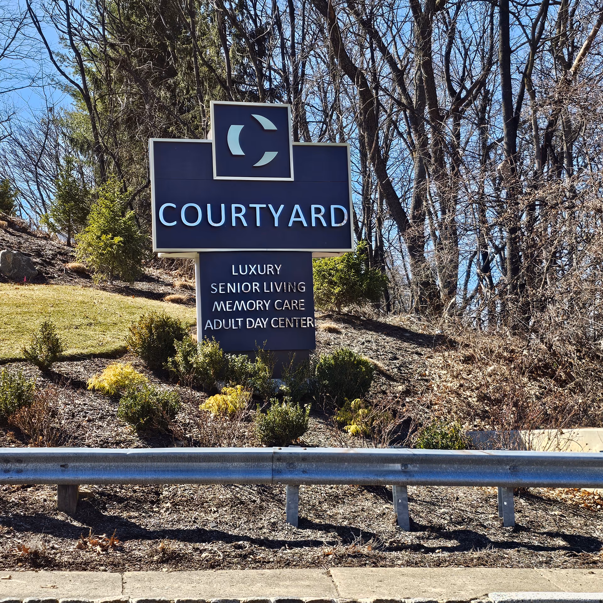 A blue Courtyard sign on a landscaped roadside with trees and a guardrail.