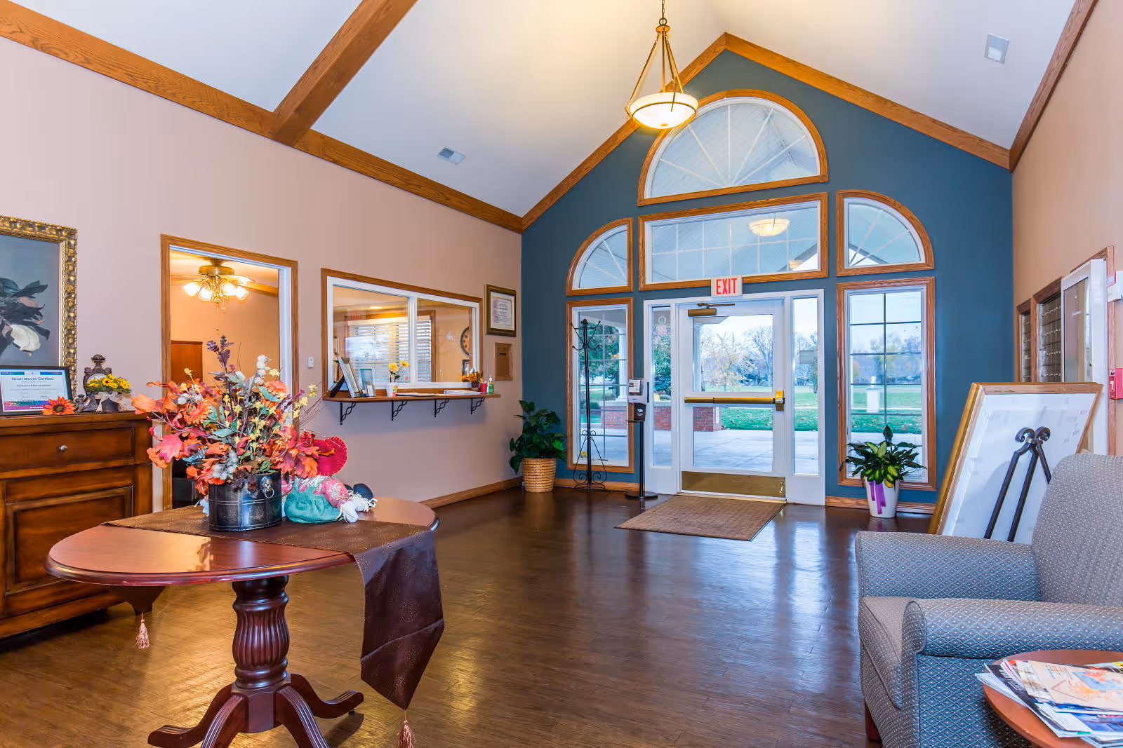 Bright lobby/entrance area with glass double doors, seating, a round table with flowers, and a reception window.