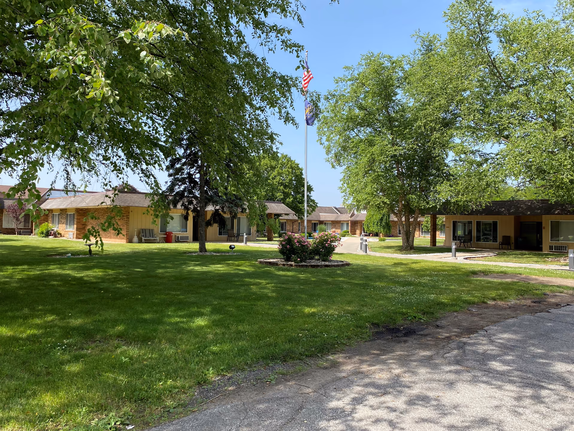 Sunlit courtyard with grassy lawn, trees, and low brick buildings around a central flagpole.