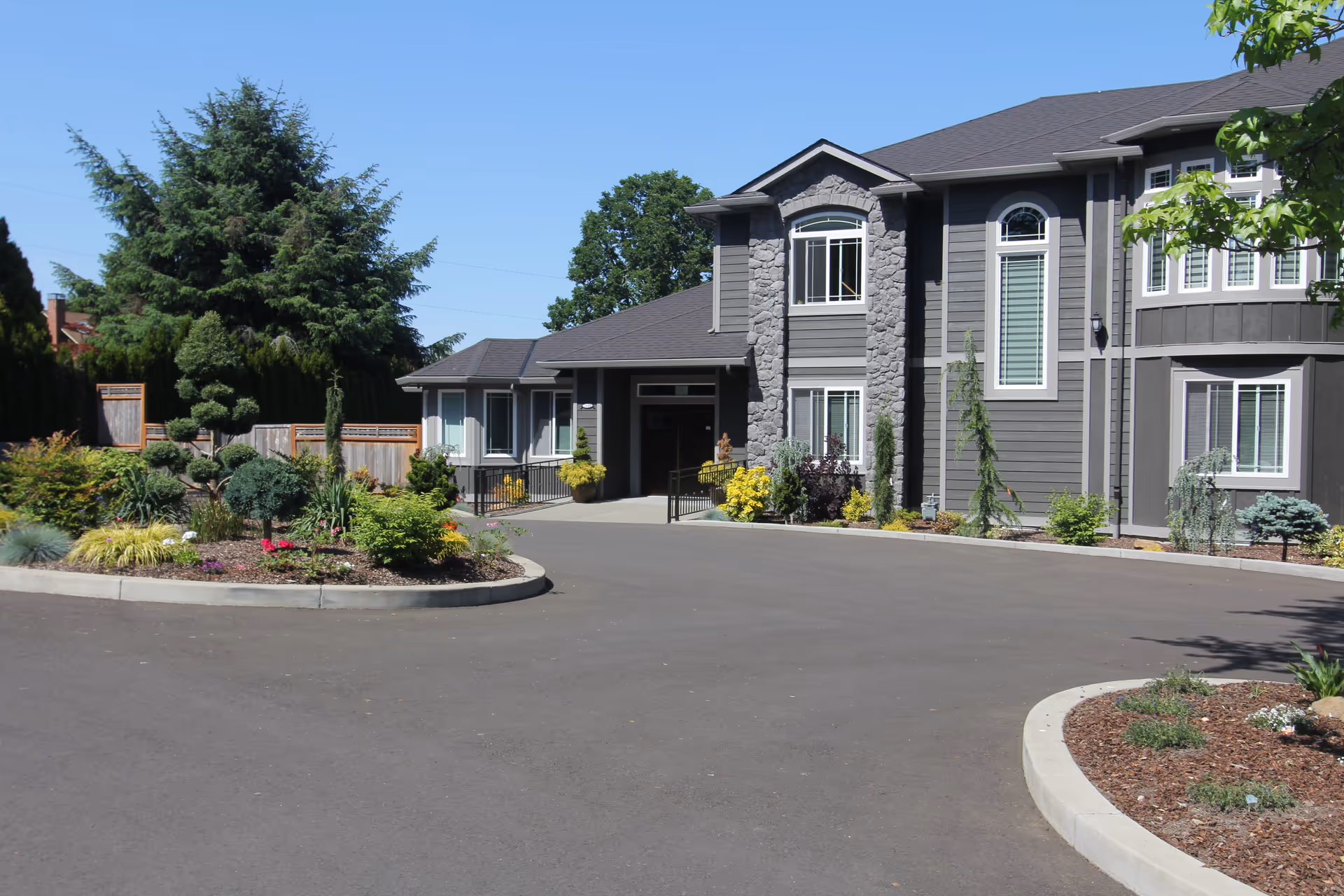 Exterior view of a senior care home building with gray siding and stone accents, surrounded by landscaped gardens and trees under a clear blue sky.