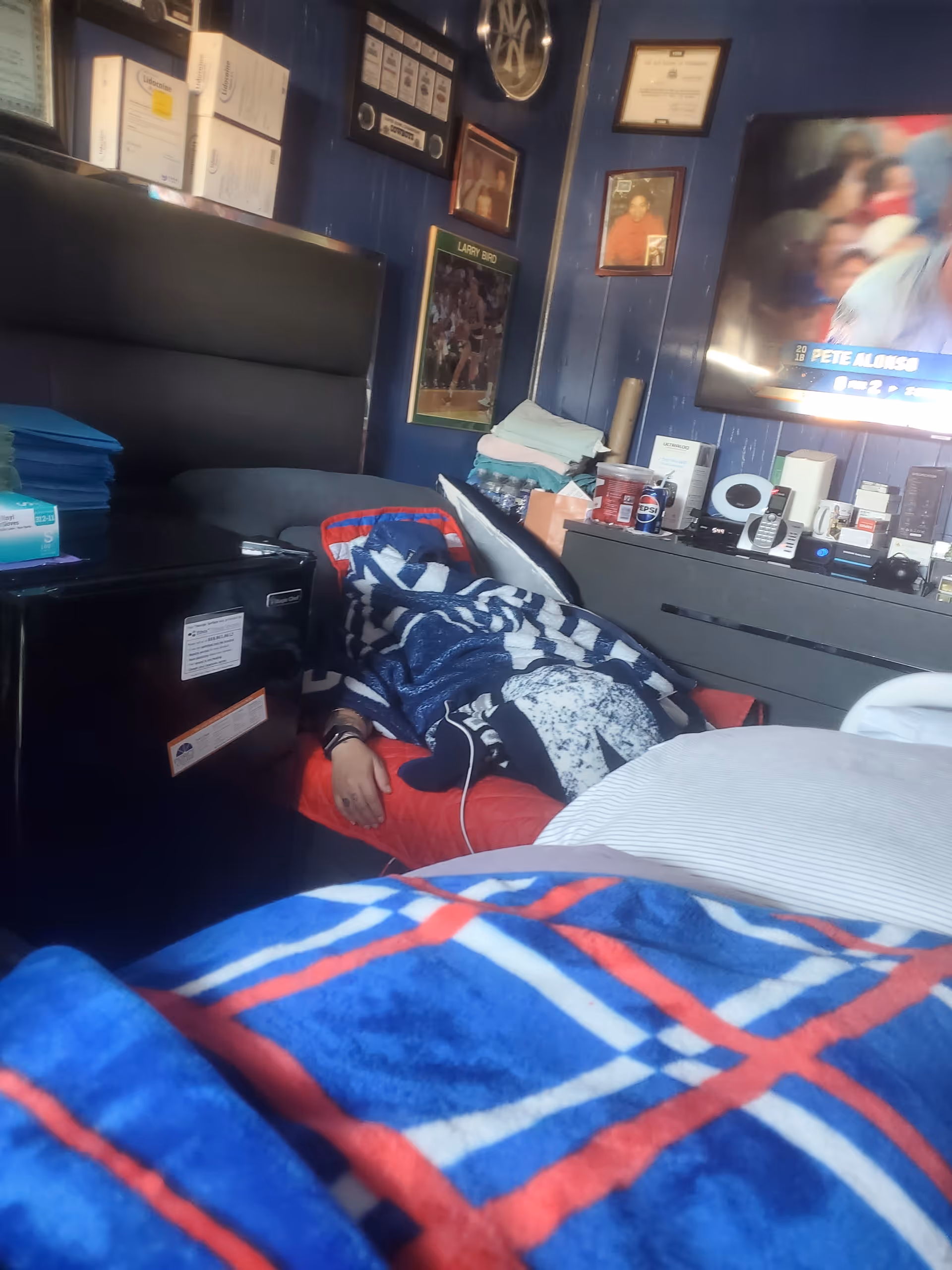 A resident partially under blankets sits in a cluttered blue-walled bedroom with a TV.