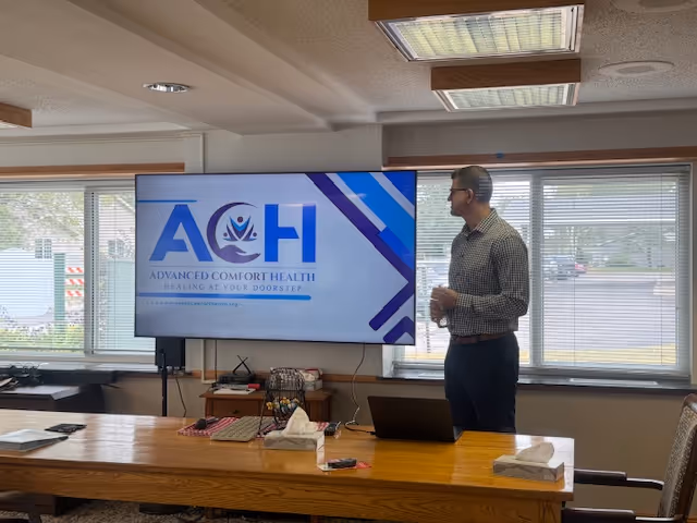 A presenter stands beside a screen displaying the Advanced Comfort Health logo in a conference room.