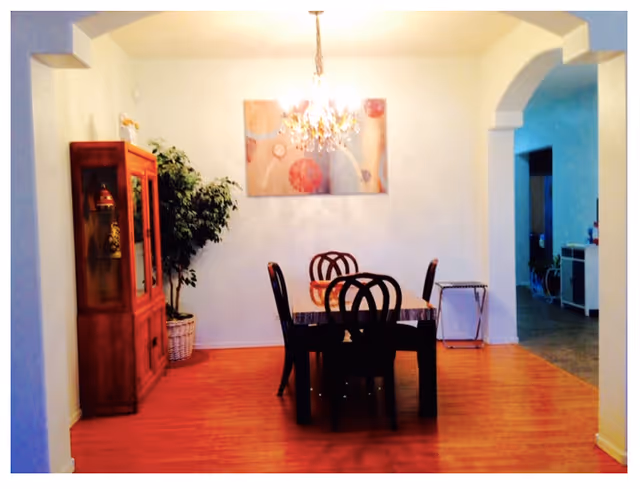 A dining room with a wooden table and six chairs under a chandelier. There is a wooden cabinet with glass doors on the left side, a potted plant next to it, and a colorful abstract painting on the wall. The room has wooden flooring and an archway leading to another area of the house.