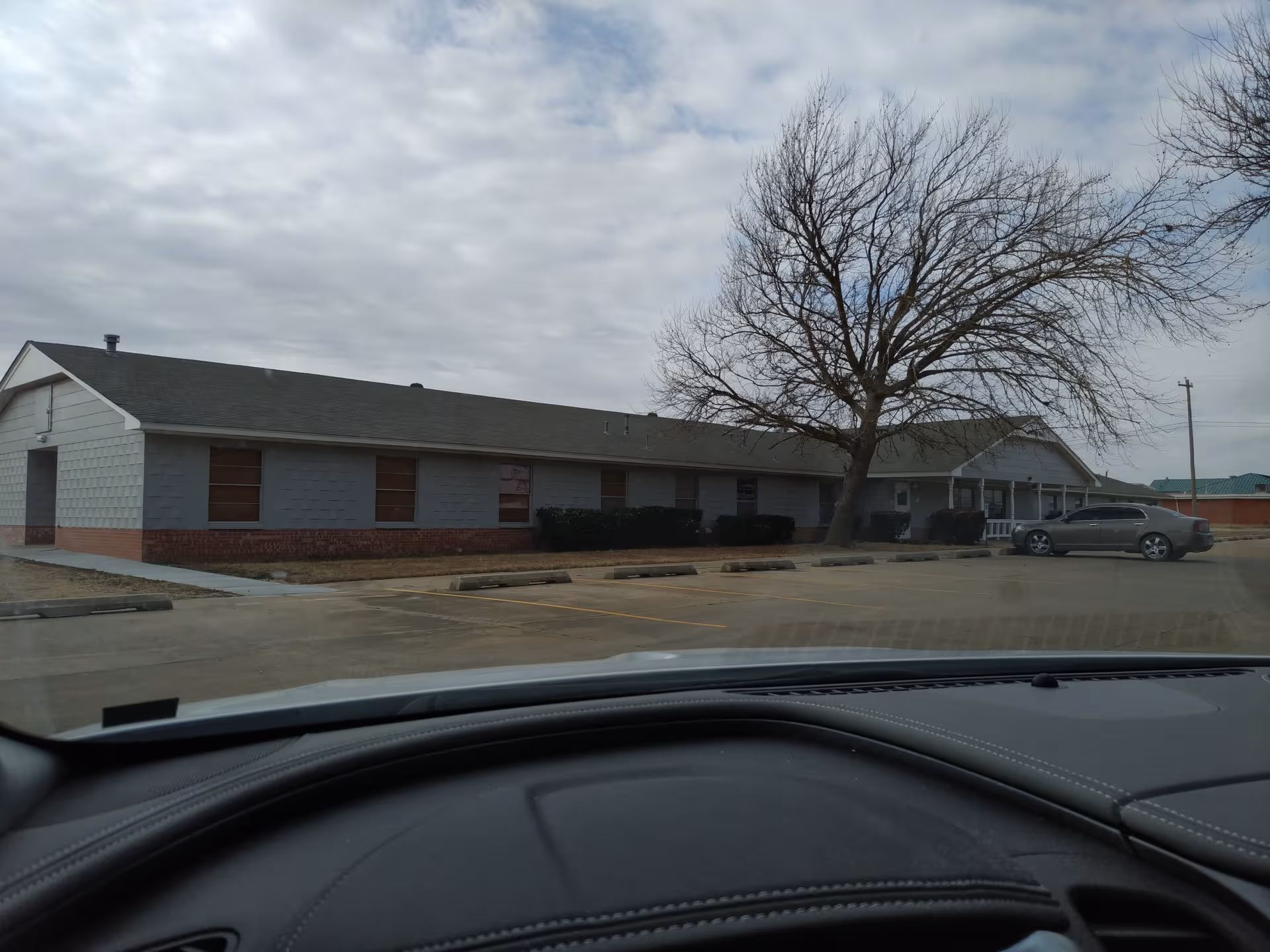Single-story assisted living building with gray siding, brick base, leafless tree, and parking.