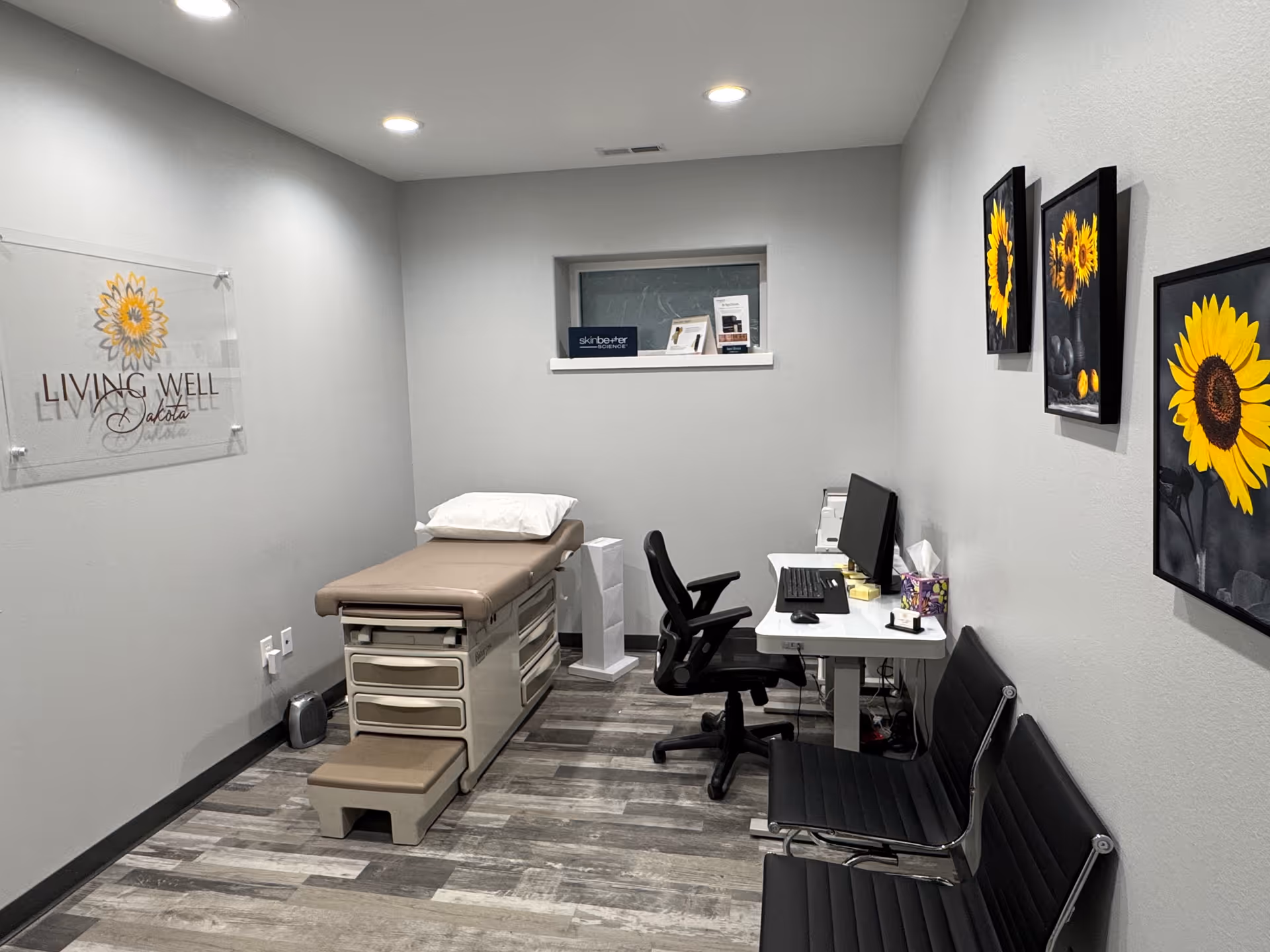 Small treatment room in Living Well Dakota with exam bed, desk, and sunflower artwork.