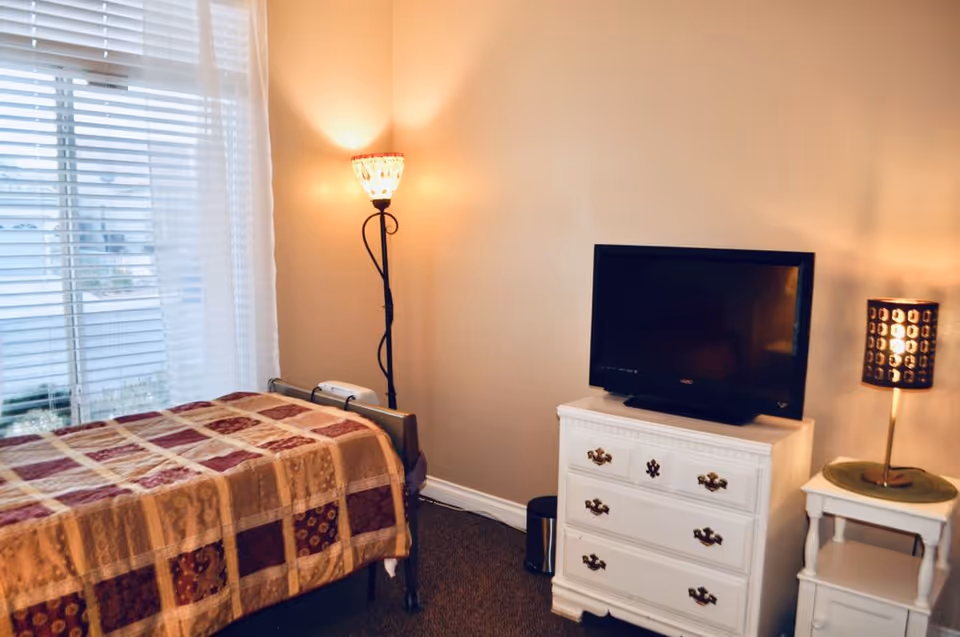 A furnished bedroom with patchwork quilt, dresser, TV, and warm lighting.