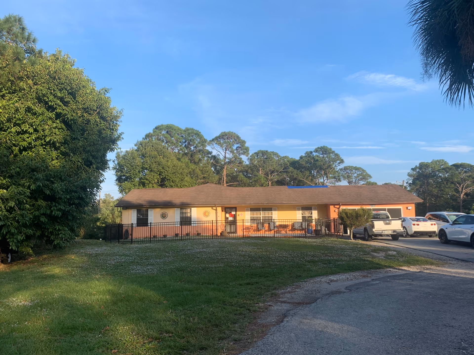 Single-story building with a brown roof and peach-colored walls surrounded by trees and a grassy lawn. Several cars are parked on the right side near the building. The sky is clear and blue.