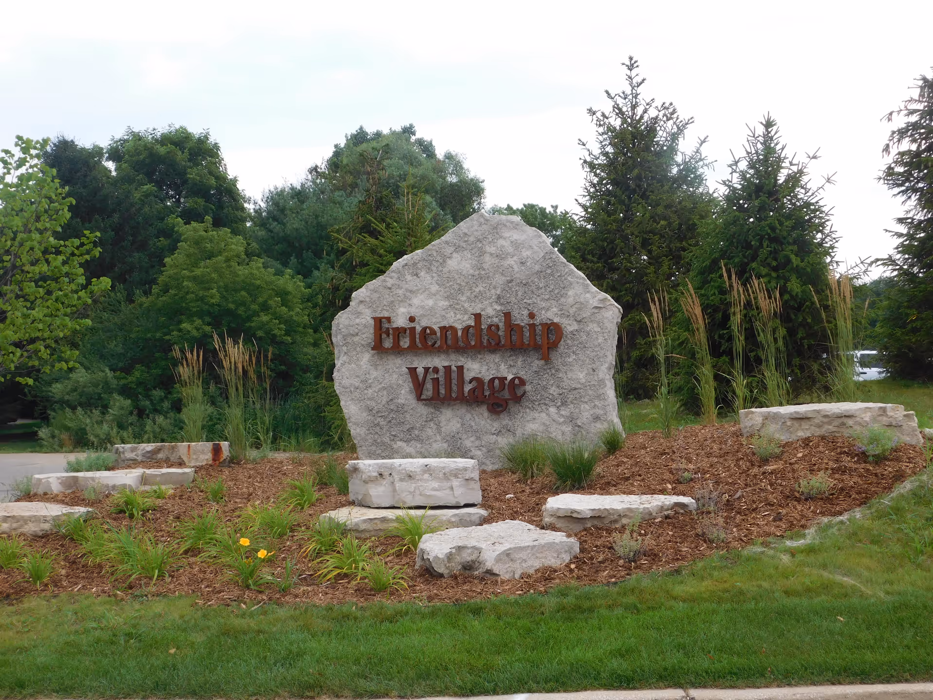 Large stone sign reading Friendship Village sits in a landscaped garden with trees.