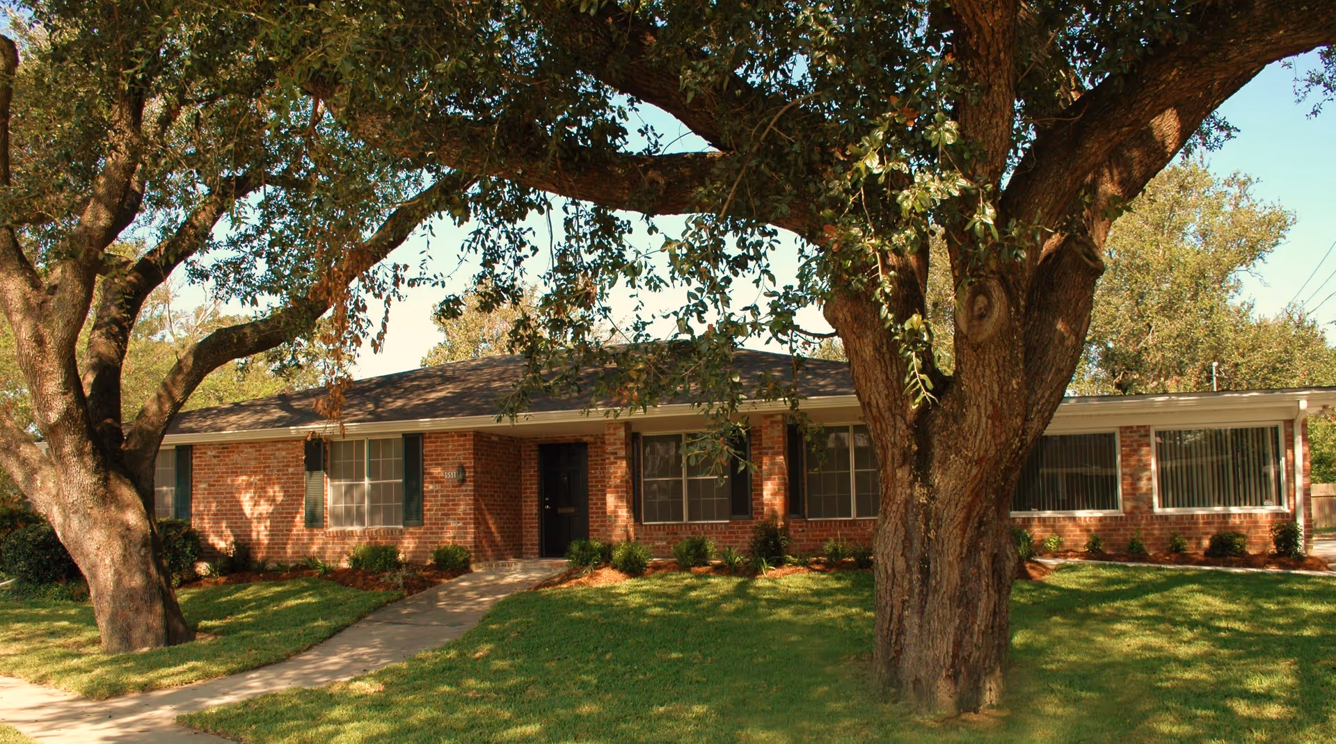 Single-story brick house with a front lawn, large overhanging trees, and a walkway leading to the entrance.