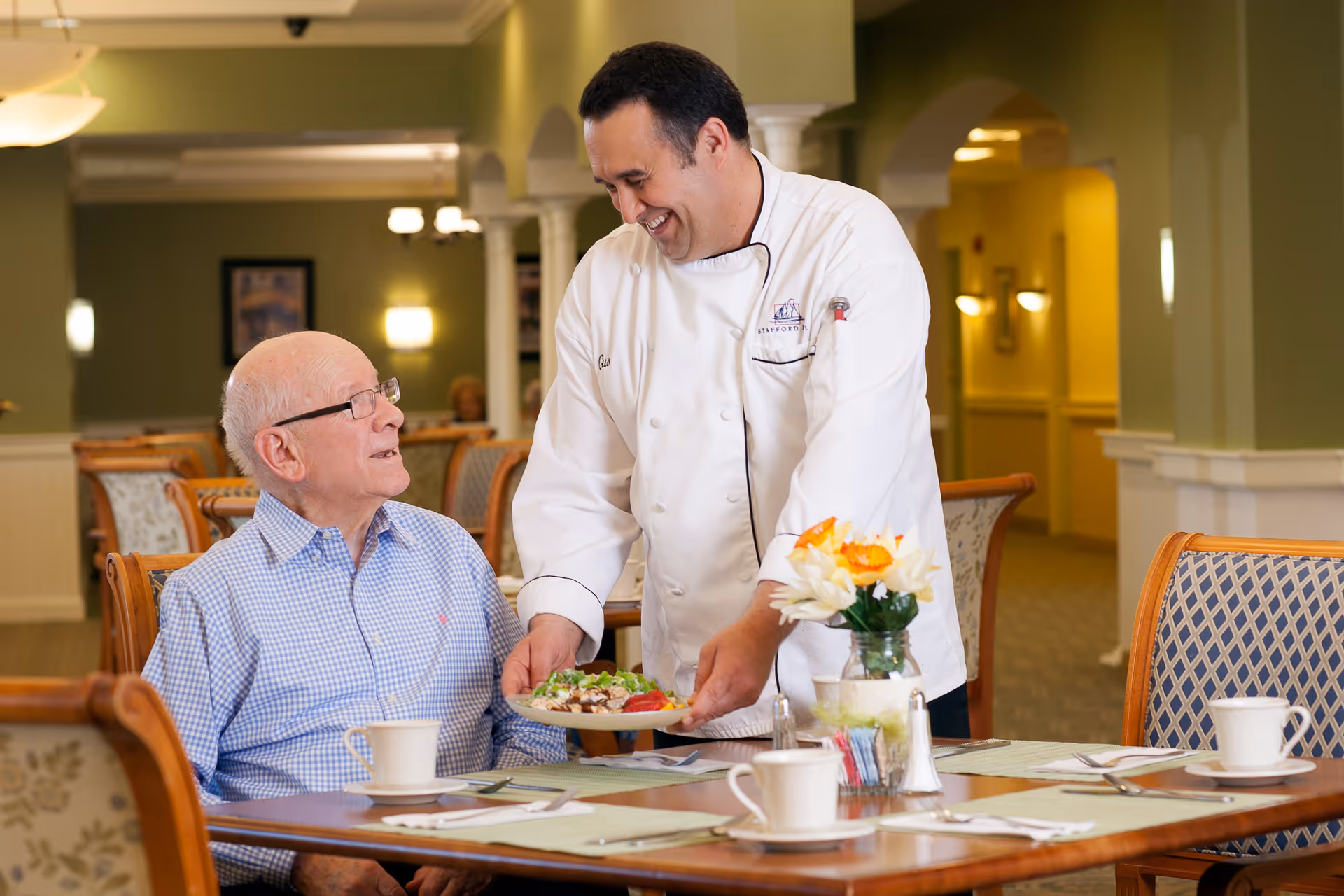 A chef in a white uniform serving a plate of food to an elderly man seated at a dining table in a well-lit dining room with green walls and wooden chairs.
