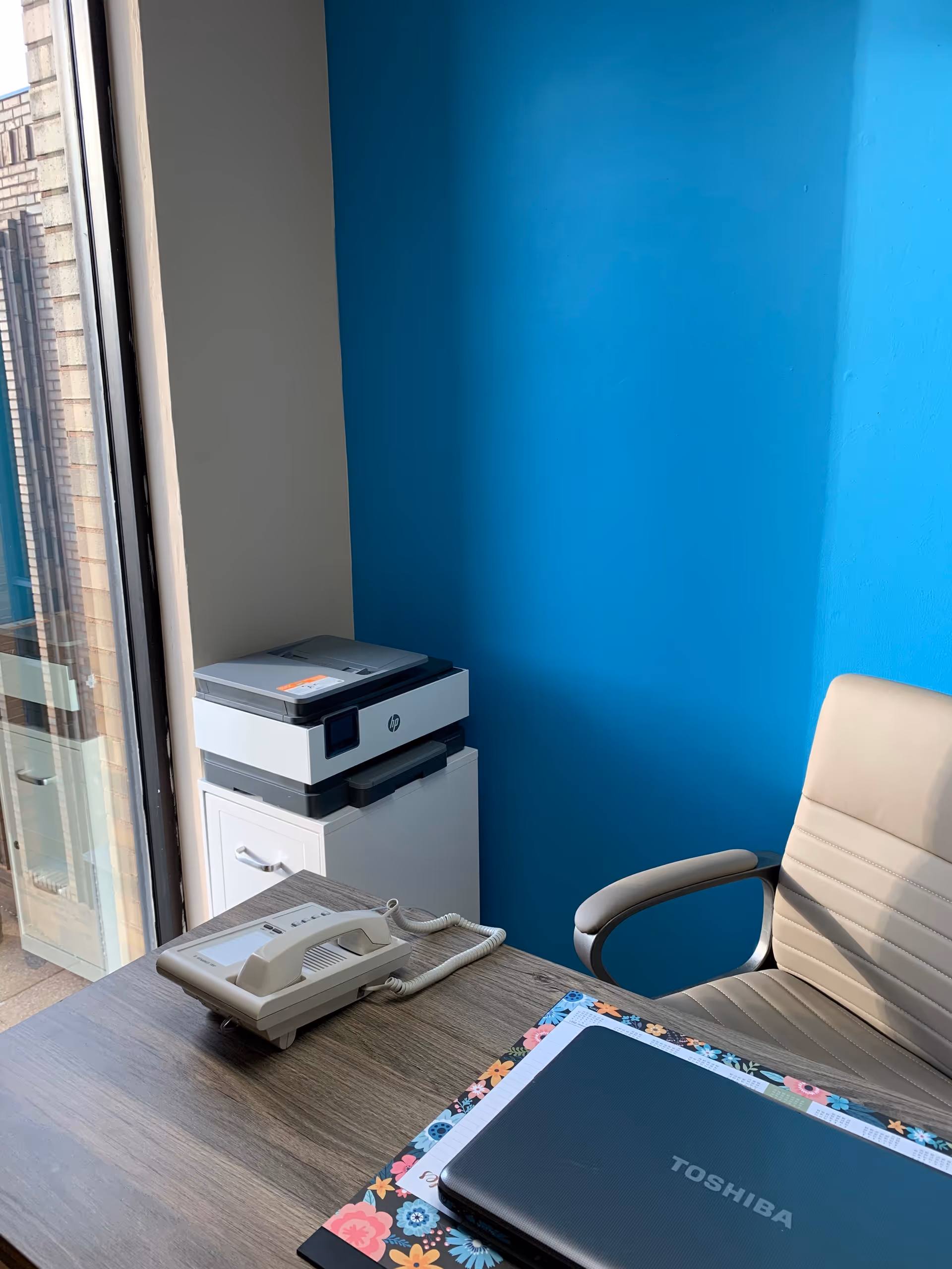 Office desk area with a Toshiba laptop, telephone, and printer against a bright blue wall.