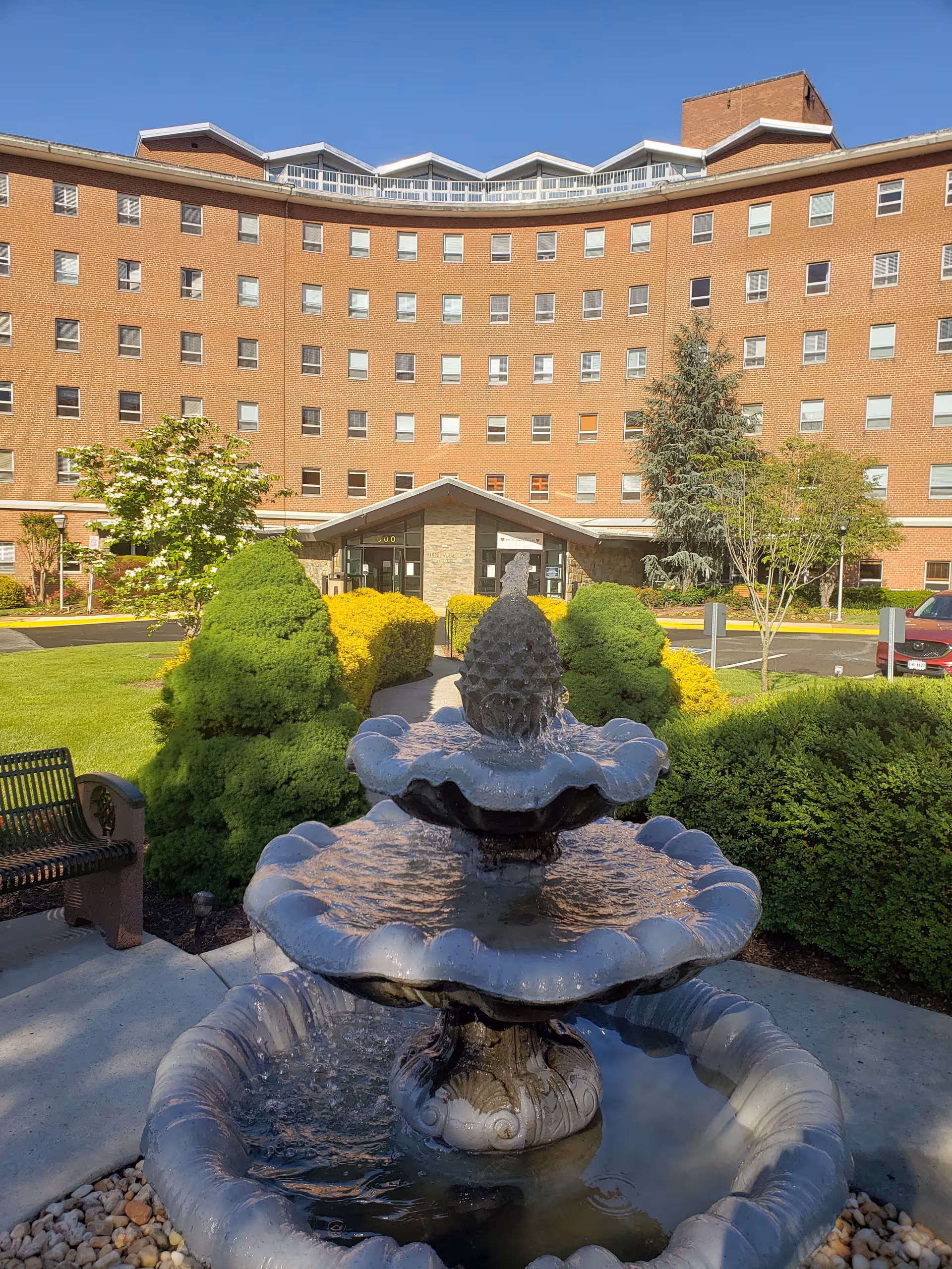 Curved brick senior living building with a tiered fountain and manicured shrubs in front.