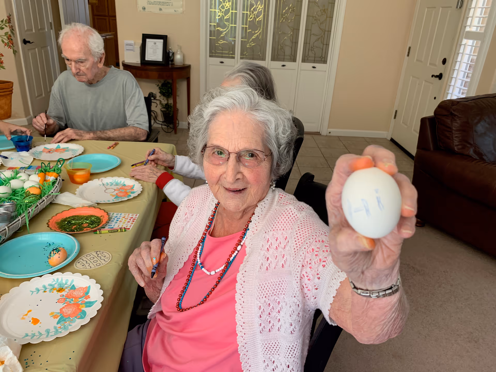 An elderly woman with gray hair and glasses is sitting at a table, smiling and holding up a decorated white egg with the word 'Hi' written on it. Around her, other elderly individuals are engaged in painting and decorating eggs. The table is covered with a yellow tablecloth and has plates, paint, and decorated eggs on it. The setting appears to be a cozy indoor common area with beige walls and a brown leather couch in the background.