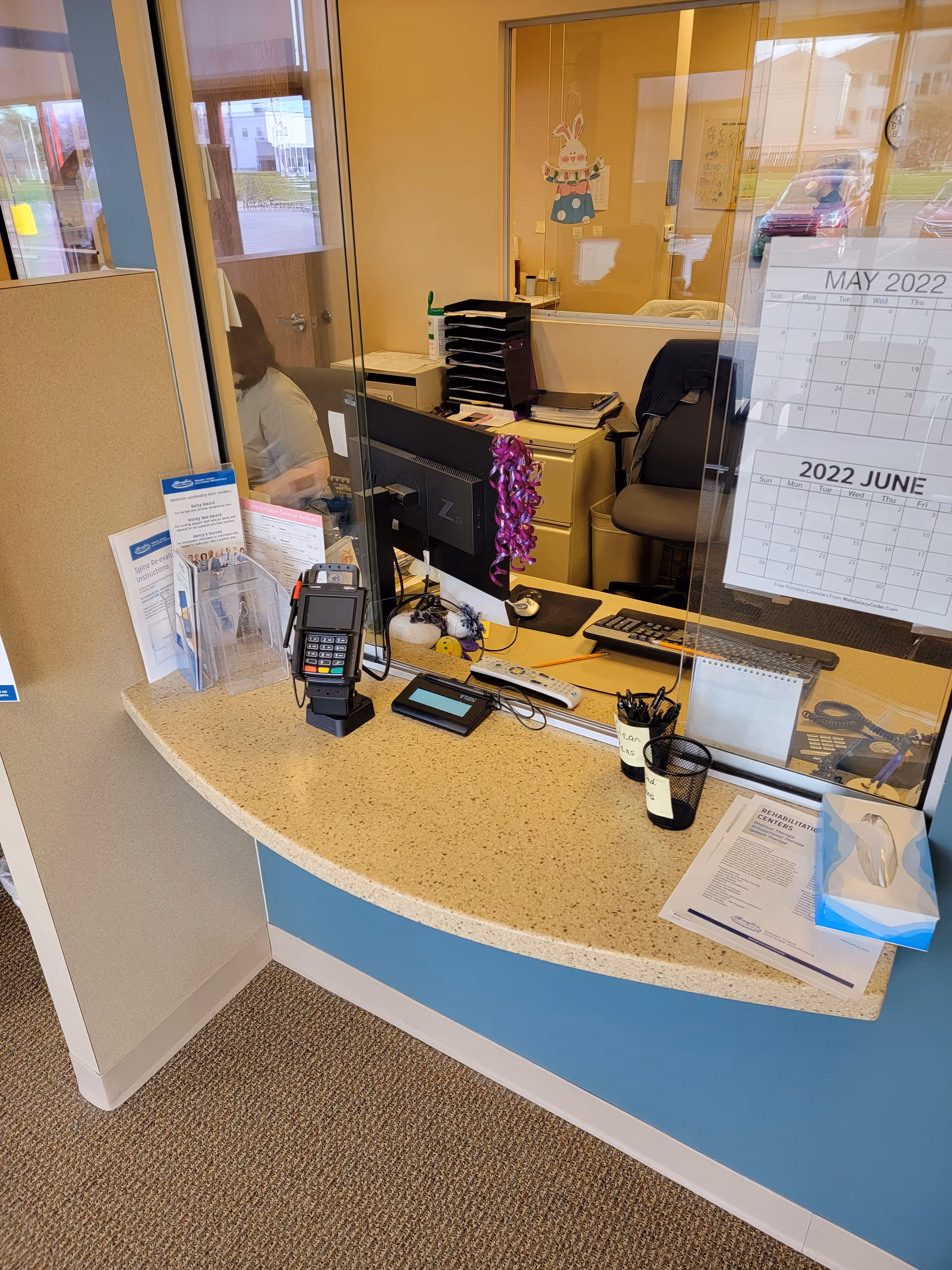 Reception desk inside a facility with calendar, payment device, paperwork, and glass partition.