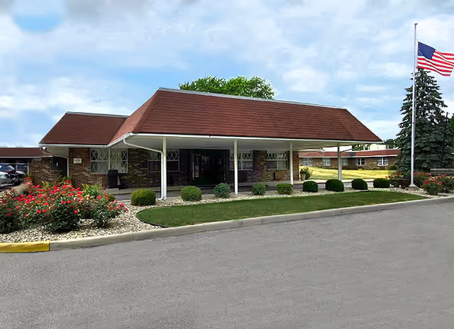 Single-story brick senior living entrance with a covered canopy, manicured landscaping, and American flag.