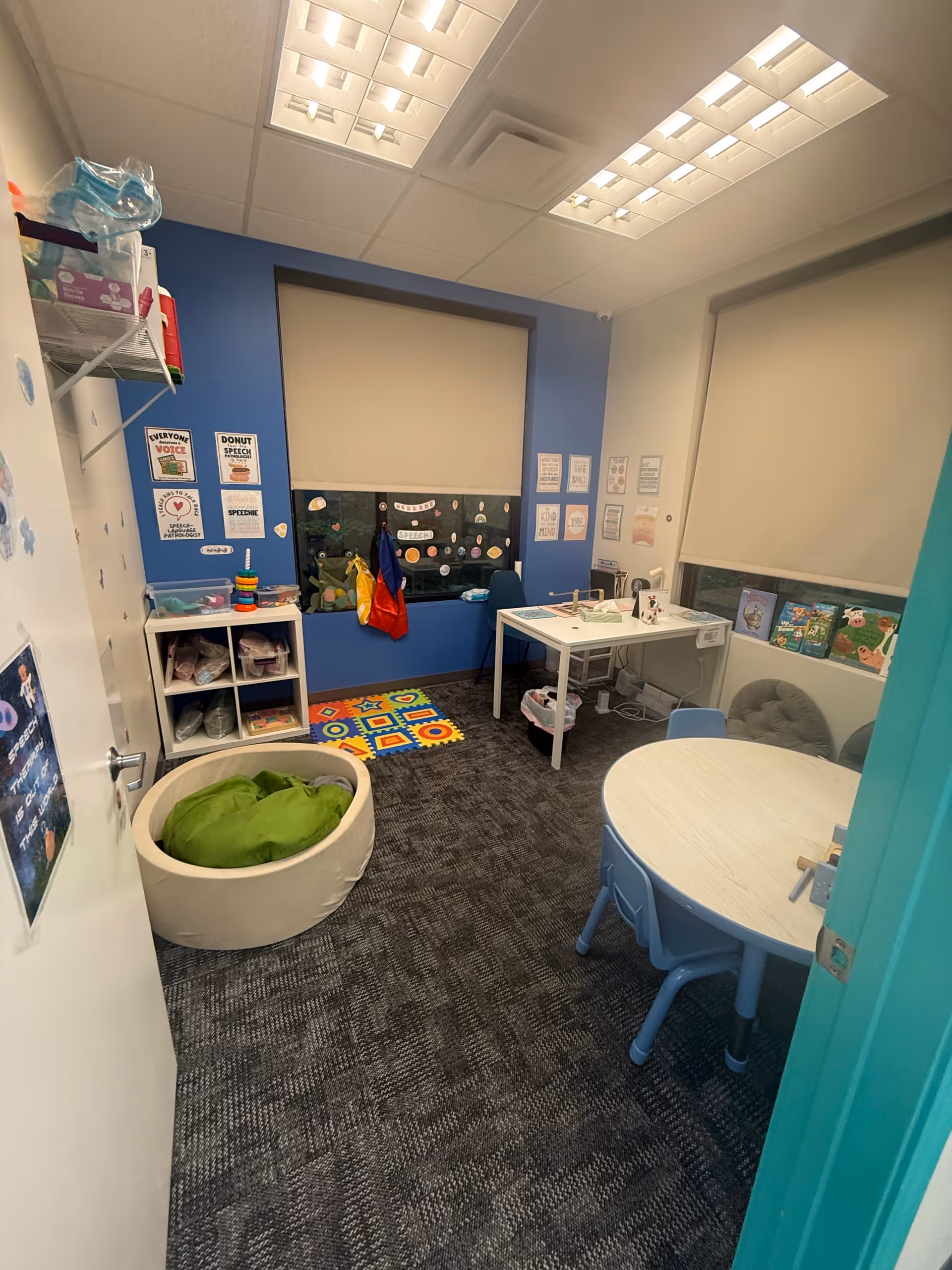 Small therapy room with blue walls, round table, chairs, desks, and educational posters.