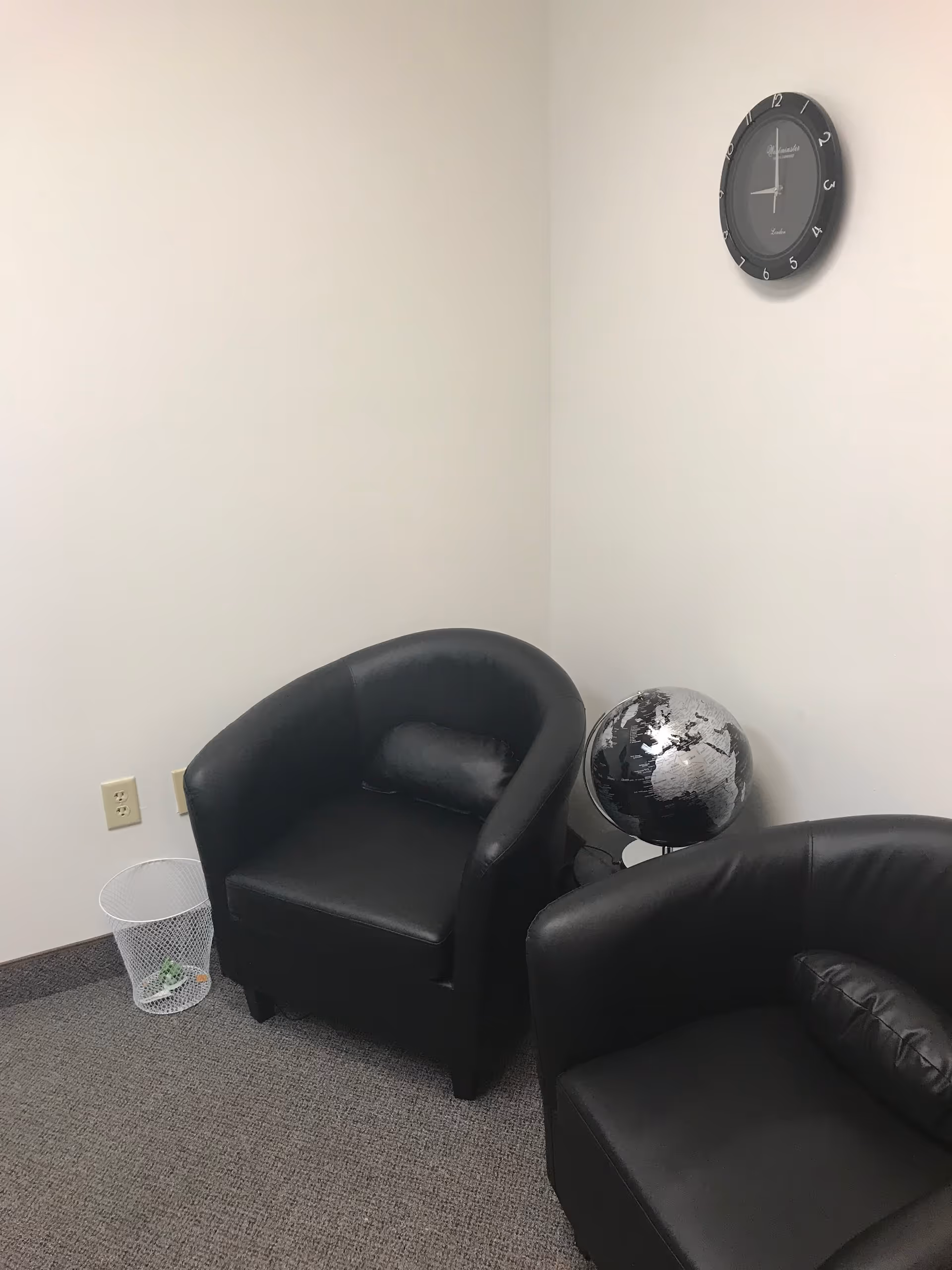 Corner of a small waiting area with two black leather chairs, a globe, and a clock.