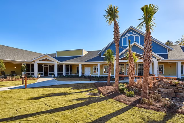 Exterior view of a single-story senior living facility building with a well-maintained lawn, palm trees, and a clear blue sky. The building features a covered entrance with white columns and a combination of yellow and blue siding.