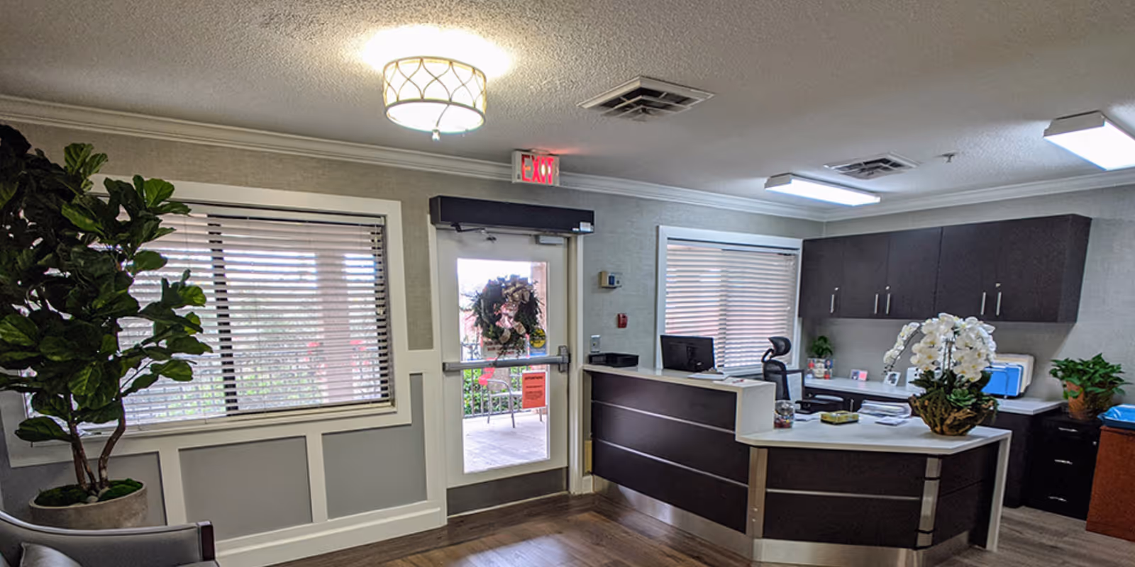 Reception area of a senior living facility with a dark wood reception desk, a computer, office chair, and decorative plants. There is a door with a wreath and an exit sign above it, windows with blinds, and ceiling lights illuminating the space.