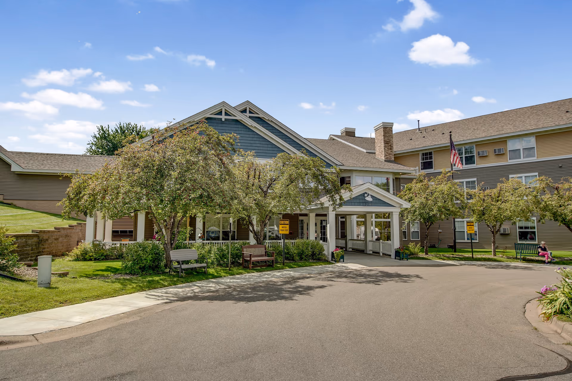 Front exterior view of Oak Ridge Assisted Living facility showing a large building with multiple windows, a covered entrance with white pillars, trees and benches in the landscaped front area, and a clear blue sky with some clouds.