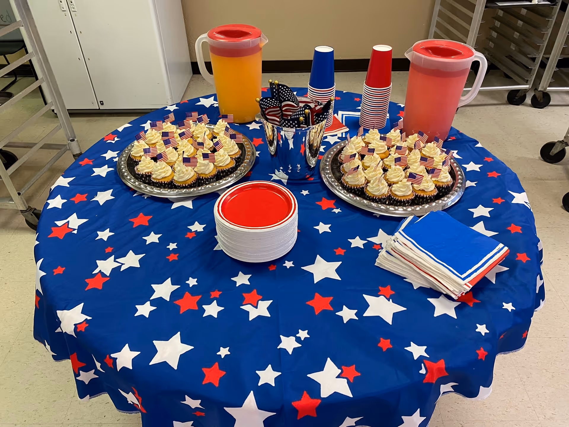Round table with star-spangled blue cloth, cupcakes, pitchers, and red napkins indoors.