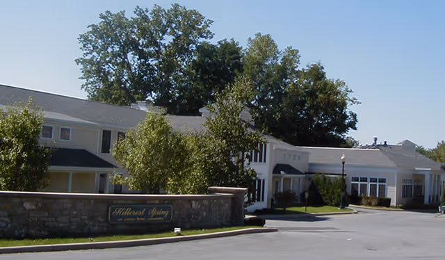 Exterior view of Hillcrest Spring Residential facility showing a large building with white siding, multiple windows, and a stone wall with a sign in front. Trees and a clear blue sky are visible in the background.