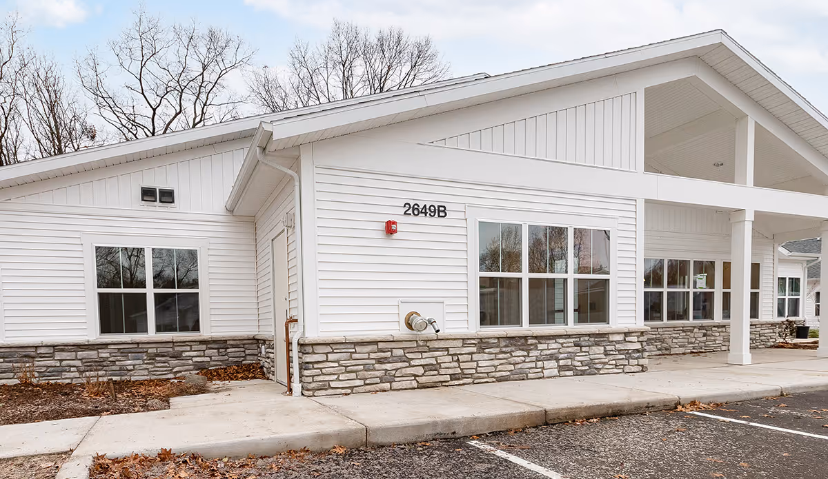 Exterior view of a single-story building with white siding and stone accents along the lower portion of the walls. The building has large windows and a covered entrance supported by white columns. The address number 2649B is visible on the wall. There are leafless trees in the background and a paved parking area in the foreground.
