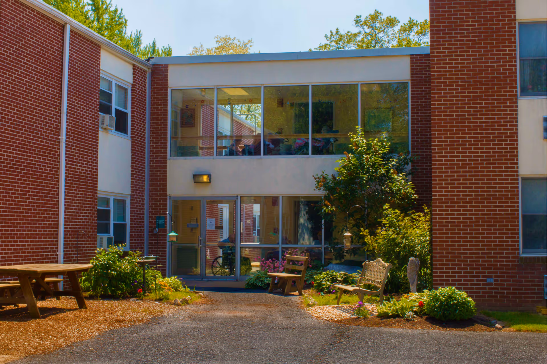 Exterior view of a senior living facility with red brick walls and large windows. There is a small garden area with plants, a wooden picnic table, a bench, and a chair near the entrance. The entrance has glass doors and the building has two stories.