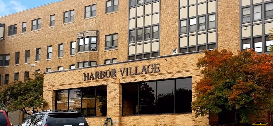 Brick senior living building with Harbor Village sign and autumn trees in front.